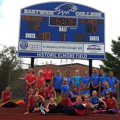 Players pose in front of the large, blue Hartwick scoreboard. The score reads two to zero Oneonta.